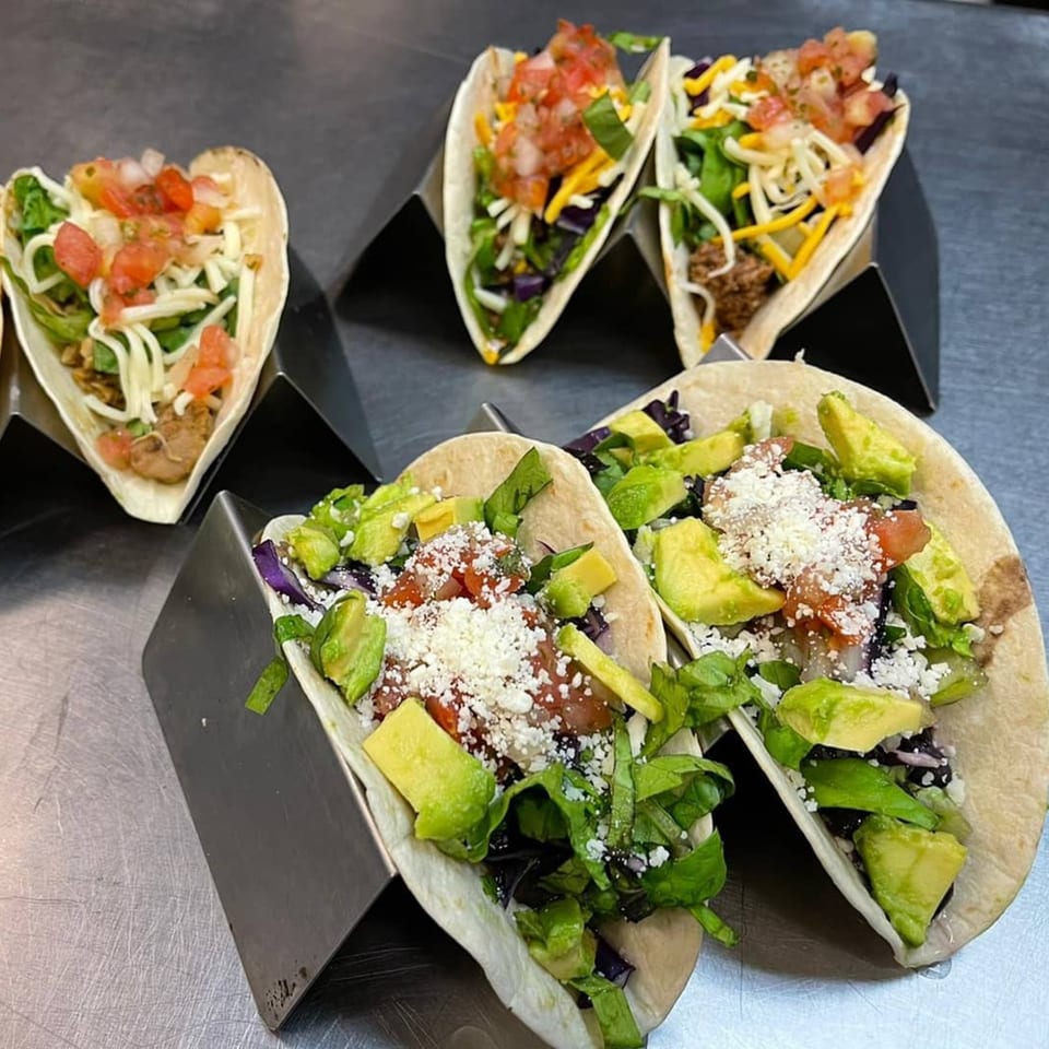 Four tacos with various fillings, including meat, cheese, lettuce, tomatoes, and avocado, on metal stands.