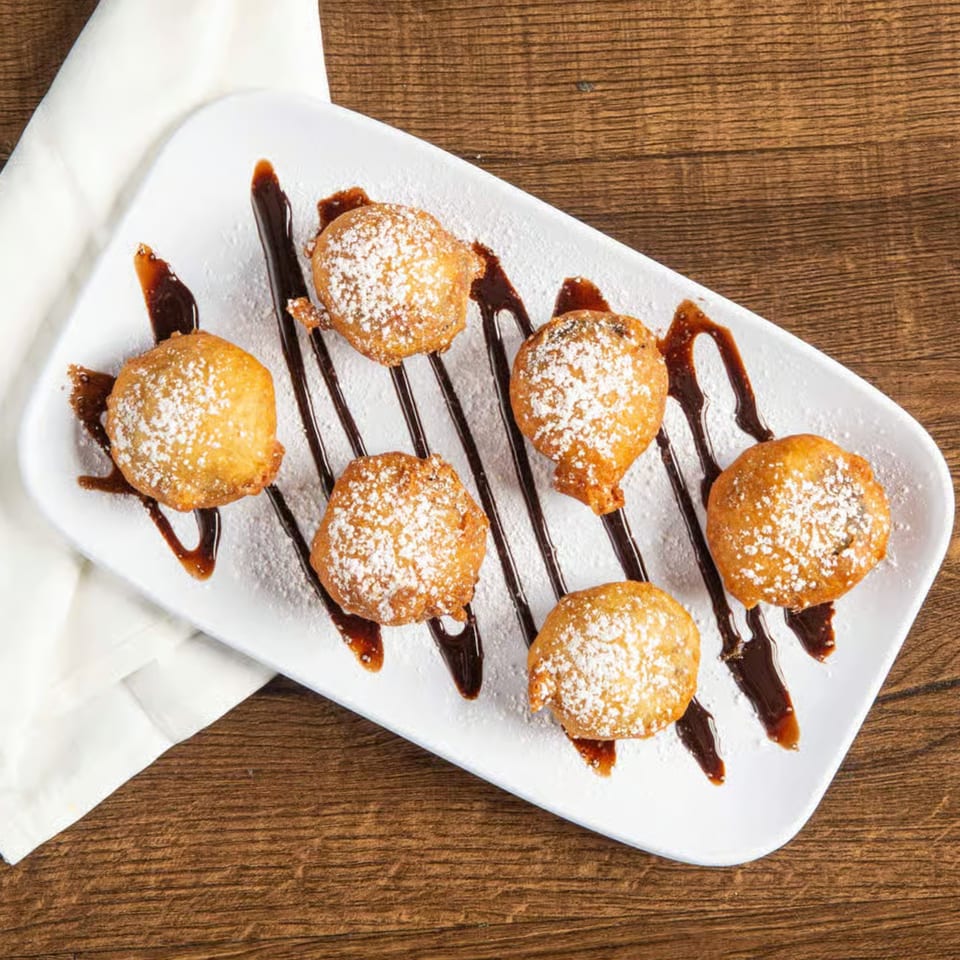 Six golden-brown fried dough balls, dusted with powdered sugar and drizzled with chocolate sauce on a white plate.