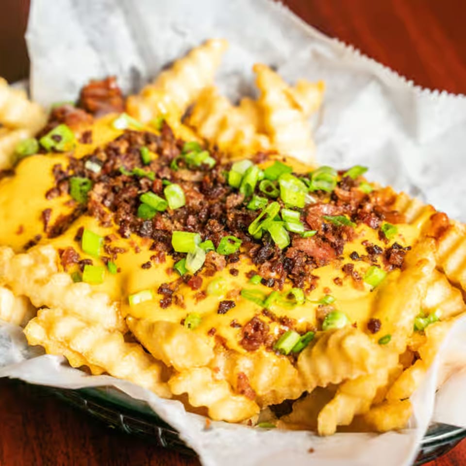 Waffle fries covered in melted cheese, crispy bacon bits, and chopped green onions in a basket.